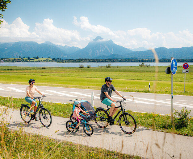 Das Bild fängt eine ruhige Szene von einer Familie mit einem Mann, einer Frau und einem kleinen Kind ein, die Fahrräder auf einem Pfad neben einer Straße fahren. Der Mann hat zudem noch einen Kinderanhänger am Fahrrad, in dem sich noch ein anderes Kleinkind befindet. Der Pfad ist von üppigem Grün umgeben. Im Hintergrund erkennt man den Hopfensee und die Berge. Der Himmel darüber ist ein klarer Blau mit fluffigen weißen Wolken. Die Radfahrer sind in legerer Kleidung und sommerlich angezogen. Sie tragen alle einen Helm. 