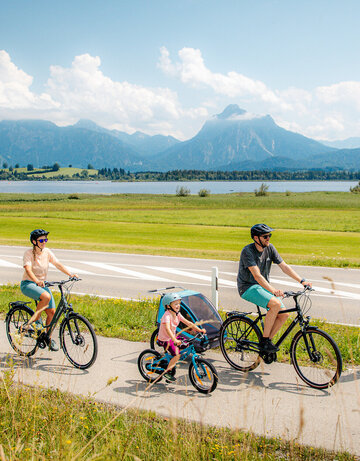 Das Bild fängt eine ruhige Szene von einer Familie mit einem Mann, einer Frau und einem kleinen Kind ein, die Fahrräder auf einem Pfad neben einer Straße fahren. Der Mann hat zudem noch einen Kinderanhänger am Fahrrad, in dem sich noch ein anderes Kleinkind befindet. Der Pfad ist von üppigem Grün umgeben. Im Hintergrund erkennt man den Hopfensee und die Berge. Der Himmel darüber ist ein klarer Blau mit fluffigen weißen Wolken. Die Radfahrer sind in legerer Kleidung und sommerlich angezogen. Sie tragen alle einen Helm. 