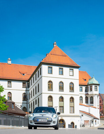 Ein silberner Mini fährt auf den Betrachter zu. Er fährt über eine Brücke. Im Hintergrund erhebt sich neben den bunten Altstadthäusern, der Gebäudekomplex des ehemaligen Benediktinerklosters mit seinen roten Dächern und unzähligen Fenstern. Der Himmel ist blau. Die Sonne scheint. 