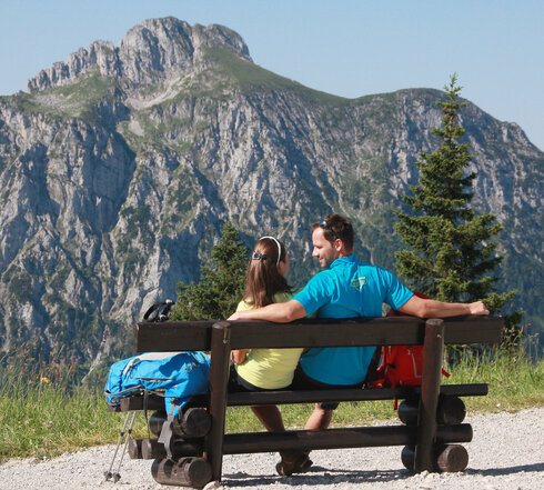 Das Bild zeigt einen Mann und eine Frau, die auf einer Holzbank sitzen. Sie tragen Wanderkleidung. Neben ihnen liegen Rucksäcke. Sie lächeln sich an. Im Hintergrund erhebt sich ein hoher Berg. 