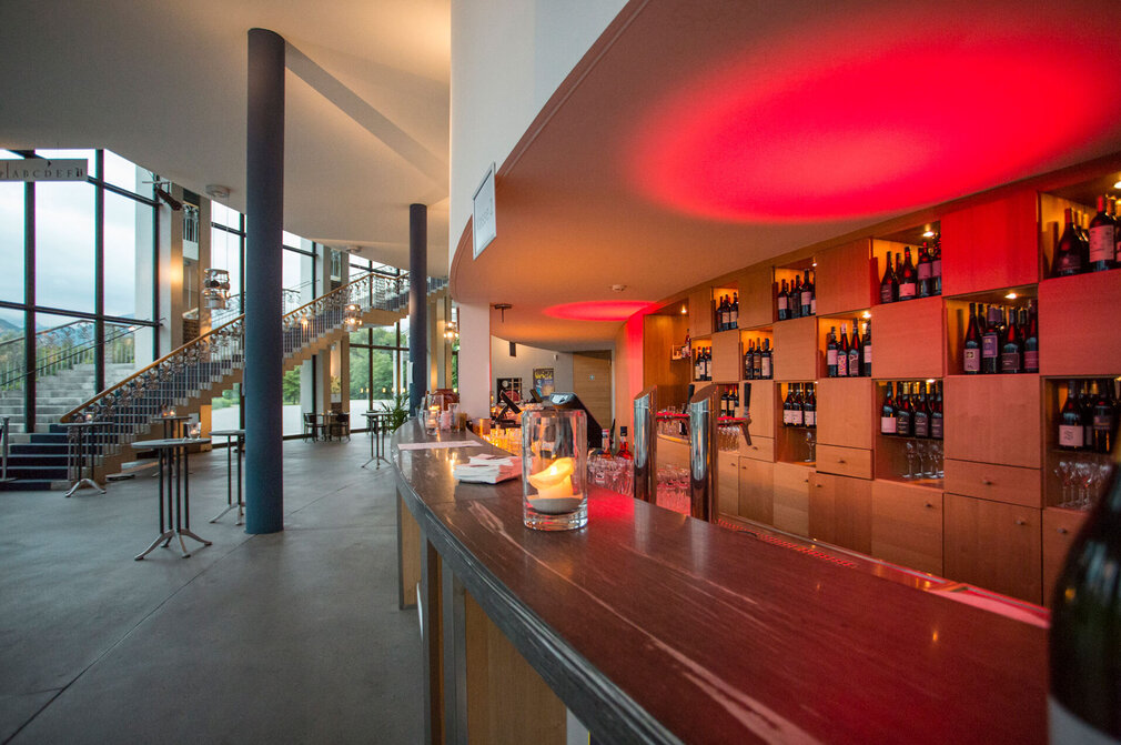 Das Foto zeigt den Tresen einer Bar in der Mitte des Bildes. Der Raum hinter dem Tresen ist in rotes Licht getaucht. Auf der Rückwand der Bar sind viele Flaschen aufgestellt. Auf dem Tresen steh ein Windlicht mit einer Kerze. Im Raum vor dem Tresen stehen Stehtische. Eine Treppe am linken Bildrand führt ins Stockwerk darüber. Durch die Fensterfront hat man einen Blick auf die Berge und Wälder. Der Himmel ist wolkenverhangen.