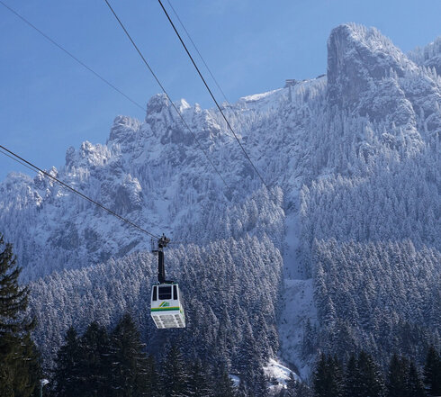 Das Foto zeigt die an einem Stahlseil hängende und talwärts oder bergwärts fahrende Kabine der Tegelbergbahn. Im Hintergrund erhebt sich der verschneite Tegelberg. Am oberen Bildrand ist die Bergstation der Bahn zu erkennen. Der Himmel ist blau. Die Sonne scheint.
