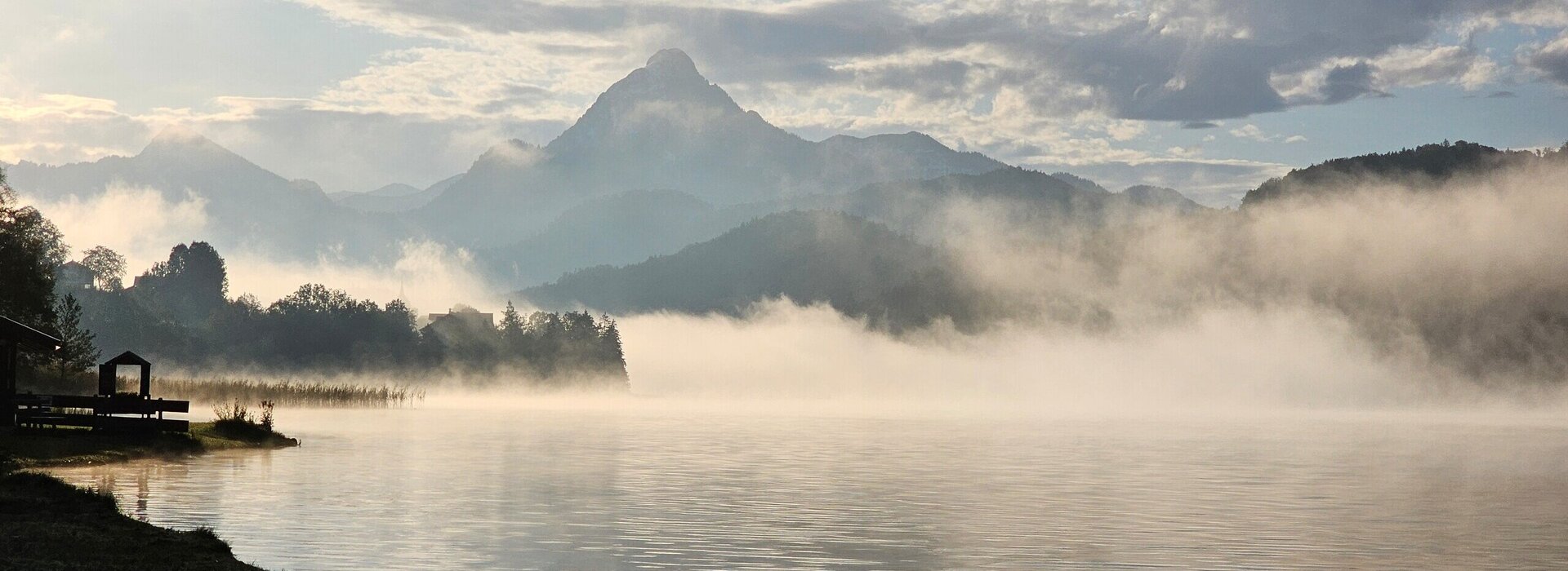 Das Bild zeigt einen See im Morgenlicht. Über den See ziehen dichte Nebelschwaden. Im Hintergrund erheben sich die Berge. Der Himmel ist blau. Die Sonne bahnt sich gerade ihren Weg.  