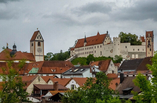 Das Foto zeigt das Hohe Schloss und die Kirche St. Mang über den Dächern von Füssen. Der Himmel ist grau und wolkenverhangen.  