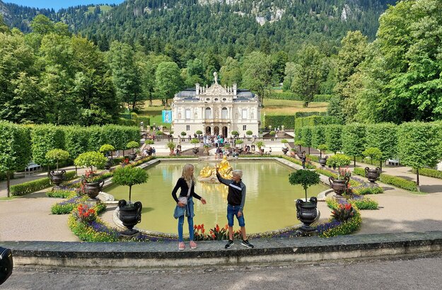  Das Bild fängt eine idyllische Szene in einem Park ein. Im Vordergrund stehen zwei Personen auf einer Steinmauer und blicken auf einen großen Teich. Der Teich ist von üppiger Vegetation umgeben, mit Bäumen und Sträuchern, die die Landschaft prägen. Ein Springbrunnen mit einer Statue befindet sich in der Mitte des Teichs und verleiht der Szene eine elegante Note. Im Hintergrund ragt ein majestätisches Burg-ähnliches Gebäude hoch, dessen Großzügigkeit durch die umgebende Vegetation betont wird. Der Himmel darüber ist ein klarer Blau, der auf einen angenehmen Tag hindeutet. Die Gesamtatmosphäre des Bildes ist eine der Ruhe und Schönheit.