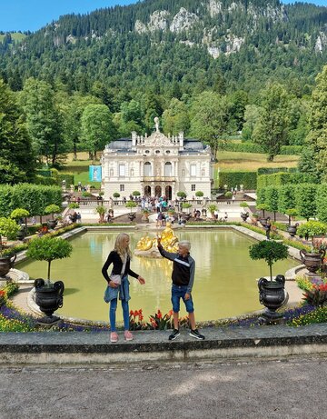  Das Bild fängt eine idyllische Szene in einem Park ein. Im Vordergrund stehen zwei Personen auf einer Steinmauer und blicken auf einen großen Teich. Der Teich ist von üppiger Vegetation umgeben, mit Bäumen und Sträuchern, die die Landschaft prägen. Ein Springbrunnen mit einer Statue befindet sich in der Mitte des Teichs und verleiht der Szene eine elegante Note. Im Hintergrund ragt ein majestätisches Burg-ähnliches Gebäude hoch, dessen Großzügigkeit durch die umgebende Vegetation betont wird. Der Himmel darüber ist ein klarer Blau, der auf einen angenehmen Tag hindeutet. Die Gesamtatmosphäre des Bildes ist eine der Ruhe und Schönheit.