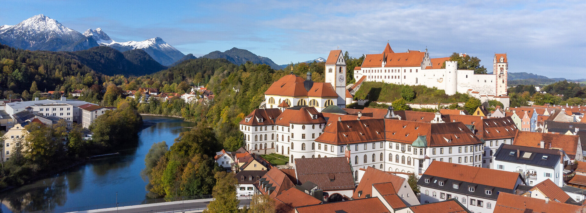 Das Foto zeigt das Hohen Schloss, dass auf einer Anhöhe über der Altstadt von Füssen thront. Direkt darunter ist der große Klosterkomplex des ehemaligen Benediktinerklosters mit seinen vielen Fenstern zu sehen. Alle Gebäude haben rote Dächer und weiße Wände. Neben den Gebäuden fließt der Lech, über den einen Brücke führt. Im Hintergrund erheben sich die Alpen, deren Gipfel schneebedeckt sind. Die Bäume haben bunte Blätter. Der Himmel ist ein wenig wolkenverhangen. 