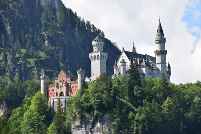 Das Foto zeigt ein Schloss mit Türmen und Zinnen. Der vordere Teil des Schlosses ist rötlich. Die Fassade des restlichen Schlosses ist weiß. Das Schloss thront auf einer Anhöhe aus schroffem Fels. Es handelt sich um das Schloss Neuschwanstein. Die Sonne scheint. Der Bergwald ringsum ist grün. Der Himmel ist blau mit wenigen Wolken durchzogen.