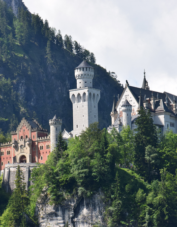 Das Foto zeigt ein Schloss mit Türmen und Zinnen. Der vordere Teil des Schlosses ist rötlich. Die Fassade des restlichen Schlosses ist weiß. Das Schloss thront auf einer Anhöhe aus schroffem Fels. Es handelt sich um das Schloss Neuschwanstein. Die Sonne scheint. Der Bergwald ringsum ist grün. Der Himmel ist blau mit wenigen Wolken durchzogen.