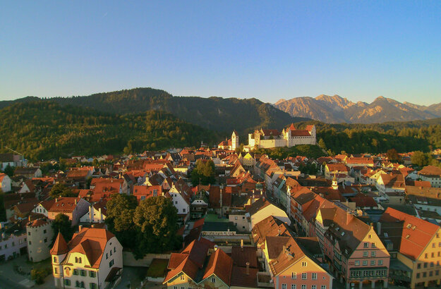 Man blickt über die roten Dächer der Stadt Füssen zu einer Anhöhe, auf der das Hohe Schloss steht. Im Hintergrund erheben sich bewaldete Hügel und hohe Berge. Der Himmel ist blau. Die Abendsonne taucht die Szene in ein warmes Licht.