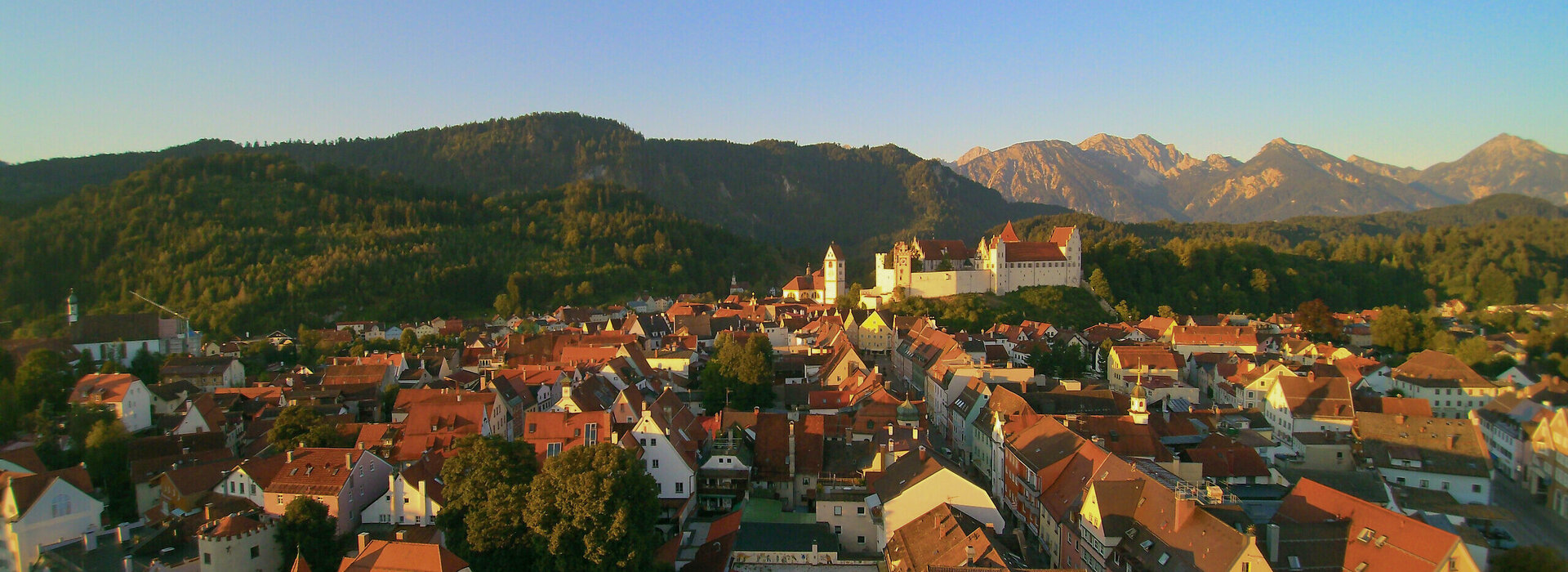 Man blickt über die roten Dächer der Stadt Füssen zu einer Anhöhe, auf der das Hohe Schloss steht. Im Hintergrund erheben sich bewaldete Hügel und hohe Berge. Der Himmel ist blau. Die Abendsonne taucht die Szene in ein warmes Licht.