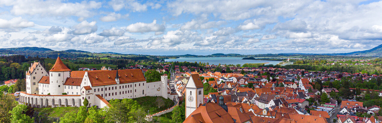 The picture offers a breathtaking view of the High Castle, the landmark of Füssen, Germany. The castle, a magnificent structure with a red roof, sits sublimely on a hill overlooking the town below. The town itself is a vibrant display of red roofs, creating an impressive contrast against the lush vegetation that surrounds it. The sky above is a clear blue, interspersed with fluffy white clouds that add to the overall serene and picturesque atmosphere of the scene. The perspective of the image is from a high view, providing a comprehensive view of the castle and the town below. The image conveys the essence of the castle and its surroundings, providing an insight into the rich history and architectural beauty of this famous landmark.