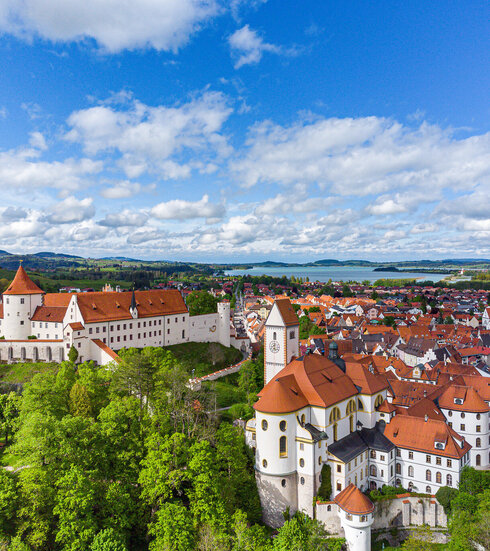 Das Bild bietet einen atemberaubenden Blick auf das Hohe Schloss, das Wahrzeichen Füssens, Deutschland. Das Schloss, eine prächtige Struktur mit einem roten Dach, thront erhaben auf einem Hügel und blickt auf die Stadt unterhalb. Die Stadt selbst ist ein lebhaftes Display von roten Dächern, die einen beeindruckenden Kontrast gegenüber der üppigen Vegetation, die sie umgibt, schaffen. Der Himmel darüber ist ein klares Blau, durchsetzt mit flauschigen weißen Wolken, die zur allgemeinen ruhigen und pittoresken Atmosphäre der Szene beitragen. Die Perspektive des Bildes ist von einer hohen Ansicht aus, was einen umfassenden Blick auf das Schloss und die Stadt darunter bietet. Das Bild vermittelt das Wesen des Schlosses und seiner Umgebung und gewährt einen Einblick in die reiche Geschichte und architektonische Schönheit dieses berühmten Wahrzeichens.