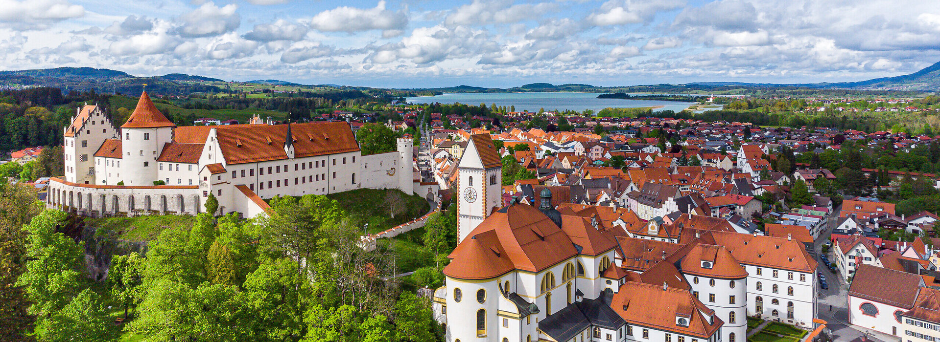 Das Bild bietet einen atemberaubenden Blick auf das Hohe Schloss, das Wahrzeichen Füssens, Deutschland. Das Schloss, eine prächtige Struktur mit einem roten Dach, thront erhaben auf einem Hügel und blickt auf die Stadt unterhalb. Die Stadt selbst ist ein lebhaftes Display von roten Dächern, die einen beeindruckenden Kontrast gegenüber der üppigen Vegetation, die sie umgibt, schaffen. Der Himmel darüber ist ein klares Blau, durchsetzt mit flauschigen weißen Wolken, die zur allgemeinen ruhigen und pittoresken Atmosphäre der Szene beitragen. Die Perspektive des Bildes ist von einer hohen Ansicht aus, was einen umfassenden Blick auf das Schloss und die Stadt darunter bietet. Das Bild vermittelt das Wesen des Schlosses und seiner Umgebung und gewährt einen Einblick in die reiche Geschichte und architektonische Schönheit dieses berühmten Wahrzeichens.
