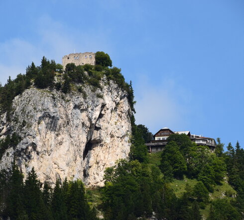 Das Bild zeigt einen atemberaubenden Ausblick auf eine Burg-ähnliche Struktur, die sich auf einem steilen Felsvorsprung befindet. Die Burg in Stein erbaut, steht majestätisch vor einem klaren blauen Himmel. Der Felsvorsprung selbst ist ein Spektakel, mit üppigem grünen Laubwerk und Büschen, die eine Naturpracht hinzufügen. Die Lage der Burg auf dem Felsvorsprung bietet einen Vorteilspunkt mit einer Panoramasicht auf das umliegende Landesinnere. Das Bild ist ein Zeugnis der architektonischen Geschicklichkeit der Vergangenheit und steht kraftvoll unter den natürlichen Elementen.