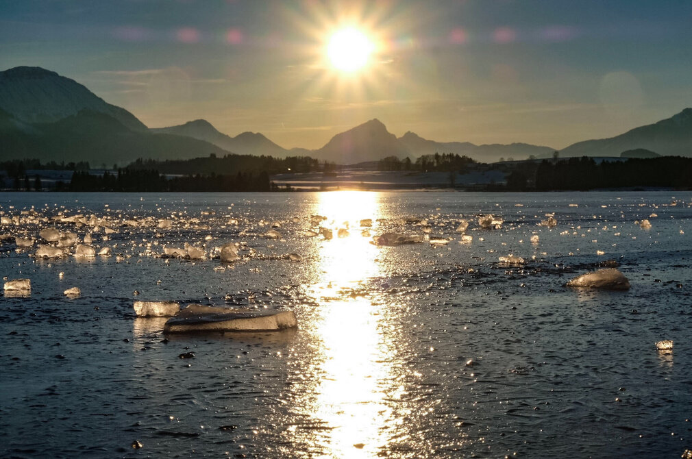 Das Bild fängt eine winterliche Szene eines Sees im Sonnenuntergang ein. Die Sonne, ein leuchtender Ball aus Licht, geht hinter einer majestätischen Bergkette unter. Der Himmel, in Orange- und Gelbtönen getaucht, spiegelt sich auf der ruhigen Wasseroberfläche des Hopfensees wider. Die Wasseroberfläche ist mit einer dicken Eisschicht bedeckt, die mit vielen Eisschollen überzogen ist. Die Berge, die sich im Hintergrund erheben, sind teilweise vom Sonnenuntergang verdeckt.