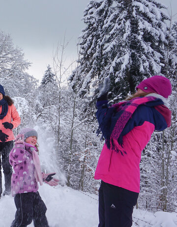 Das Bild fängt eine fröhliche Winterszene in einem schneebedeckten Wald ein. Eine Gruppe von Menschen, die in bunten Winterbekleidungen gekleidet sind, genießt den Schnee. Einige von ihnen werfen Schneebälle, was eine spielerische Atmosphäre schafft. Die Bäume im Hintergrund sind mit Schnee bedeckt, was zur winterlichen Atmosphäre beiträgt. Der Himmel über ihnen ist wolkenverhangen, was auf einen kalten und verschneiten Tag hinweist. Die Menschen stehen auf einem schneebedeckten Weg, der durch den Wald führt. Die gesamte Szene vermittelt ein Gefühl von Spaß und Kameradschaft, da die Gruppe von Freunden oder Familienmitgliedern Zeit miteinander in den Schnee verbringt.