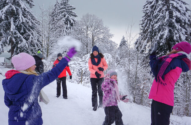 Das Bild fängt eine fröhliche Winterszene in einem schneebedeckten Wald ein. Eine Gruppe von Menschen, die in bunten Winterbekleidungen gekleidet sind, genießt den Schnee. Einige von ihnen werfen Schneebälle, was eine spielerische Atmosphäre schafft. Die Bäume im Hintergrund sind mit Schnee bedeckt, was zur winterlichen Atmosphäre beiträgt. Der Himmel über ihnen ist wolkenverhangen, was auf einen kalten und verschneiten Tag hinweist. Die Menschen stehen auf einem schneebedeckten Weg, der durch den Wald führt. Die gesamte Szene vermittelt ein Gefühl von Spaß und Kameradschaft, da die Gruppe von Freunden oder Familienmitgliedern Zeit miteinander in den Schnee verbringt.