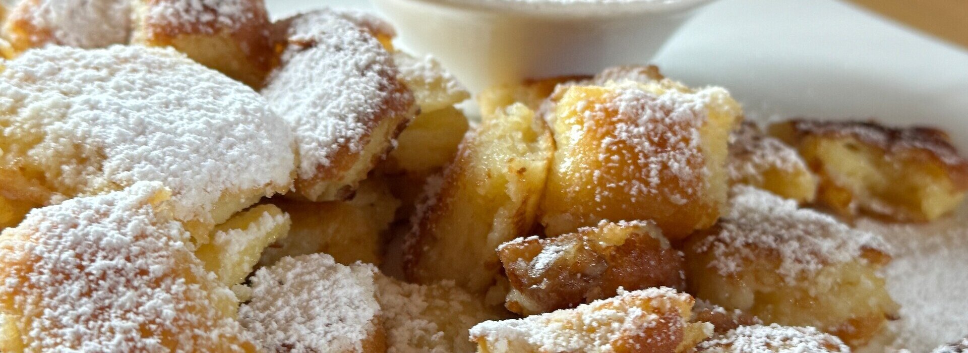 Das Bild zeigt eine verführerische Dessert-Szene. Im Zentrum des Bildes ist ein weißer Teller zu sehen, der eine großzügige Portion goldbrauner Pfannkuchen hält. Die Pfannkuchen sind mit Puderzucker bestreut, was ihnen eine süße Note verleiht.  Zur Seite des Tellers hin steht ein kleines weißes Schälchen, gefüllt mit einer cremigen gelben Sauce. Die Sauce scheint eine Art Custard oder Sahne zu sein und ist bereit, über die Pfannkuchen geträufelt zu werden.  Der Teller steht auf einem hölzernen Tisch, was dem Gesamtaussehen einen rustikalen Charme verleiht. Im Hintergrund ist eine Person, teilweise sichtbar, was darauf schließen lässt, dass es sich um ein gemütliches Frühstück oder Brunch handeln könnte. Die Person scheint das Essen zu genießen, was eine Wärme und Geborgenheit in die Szene bringt.
