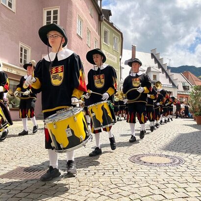 Das Bild zeigt einen Festzug. Eine Gruppe historisch gekleideter Personen mit gelb-schwarzen Trommeln laufen am Betrachter vorbei. Alle Personen haben Trommelstöcke in den Händen. Sie laufen durch eine Straße in Füssen. Im Hintergrund sind bunte Häuserfassaden zu erkennen. Viele Personen stehen am Straßenrand und beobachten den Festzug. Der Himmel ist blau mit weißen Wolken. Die Sonne scheint.