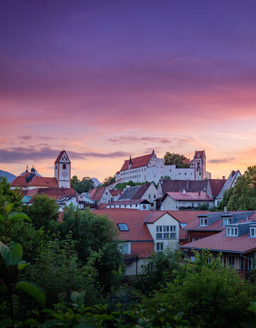  Das Bild fängt eine friedliche Sonnenuntergangsszene in Füssen ein. Der Himmel, der in Farbtönen von Lila und Pink gemalt ist, dient als schöne Kulisse für die Altstadt. Das Bild selbst ist ein charmanter Mix aus weißen und roten Gebäuden, deren Dächer eine Farbtupfer in die Szene bringen. Die allgemeine Atmosphäre des Bildes ist friedvoll und idyllisch und fängt das Wesen eines ruhigen kleinen Ortes in der Abenddämmerung ein.