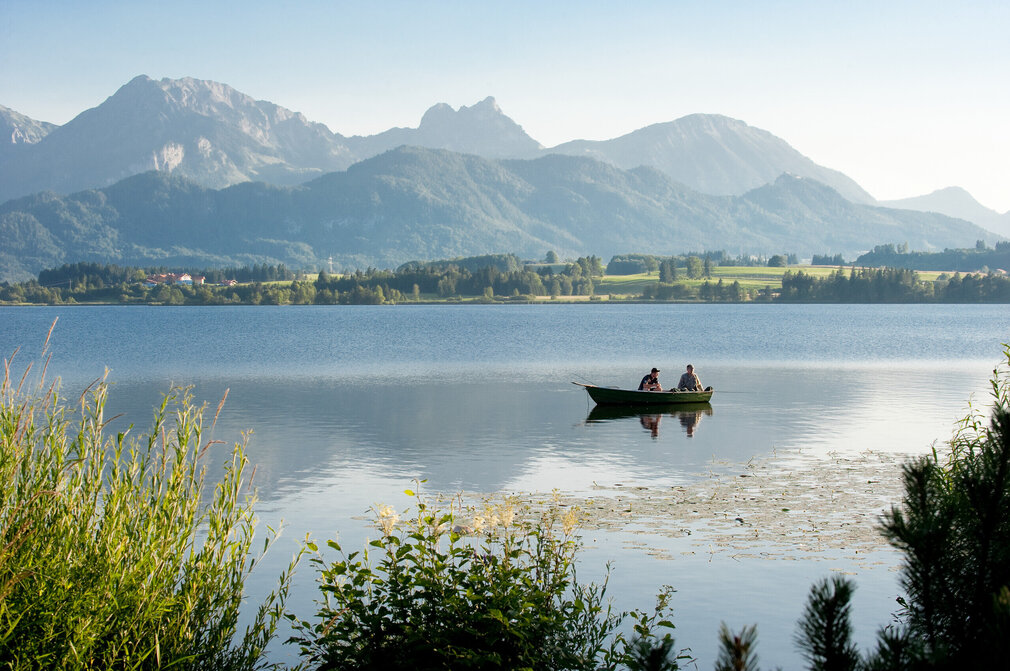 Das Bild fängt eine friedliche Szene von zwei Menschen in einem kleinen Ruderboot ein, das sanft auf dem ruhigen Hopfensee gleitet. Das Boot, in einem lebendigen Grünton gestrichen, ist im Zentrum des Bildes positioniert. Die Personen im Boot scheinen einen friedvollen Moment zu genießen, vielleicht die atemberaubende Aussicht der majestätischen Berge zu bewundern, die die Kulisse des Bildes bilden. Die Berge erheben sich majestätisch gegen den klaren Himmel und tragen zur Ruhe des Szenarios bei. Der See, ein Spiegel des Himmels, reflektiert die Schönheit der Umgebung und verstärkt die allgemeine friedliche Atmosphäre des Bildes.