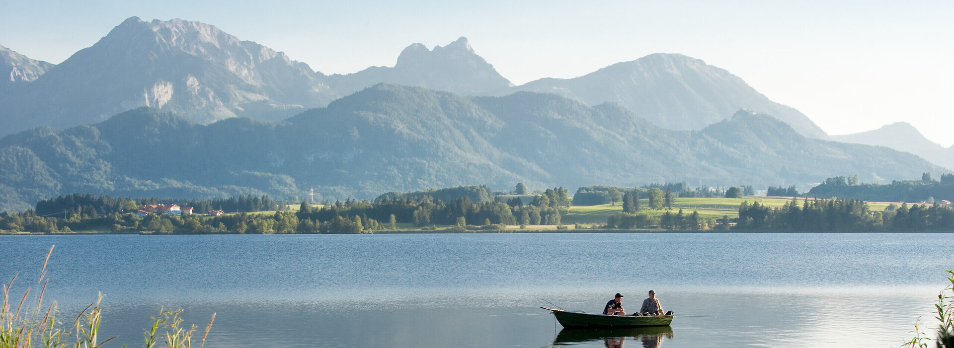 Das Bild fängt eine friedliche Szene von zwei Menschen in einem kleinen Ruderboot ein, das sanft auf dem ruhigen Hopfensee gleitet. Das Boot, in einem lebendigen Grünton gestrichen, ist im Zentrum des Bildes positioniert. Die Personen im Boot scheinen einen friedvollen Moment zu genießen, vielleicht die atemberaubende Aussicht der majestätischen Berge zu bewundern, die die Kulisse des Bildes bilden. Die Berge erheben sich majestätisch gegen den klaren Himmel und tragen zur Ruhe des Szenarios bei. Der See, ein Spiegel des Himmels, reflektiert die Schönheit der Umgebung und verstärkt die allgemeine friedliche Atmosphäre des Bildes.