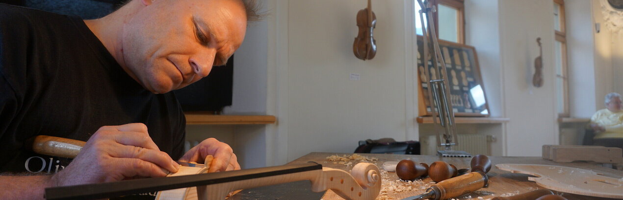 Man blickt auf einen Mann, der mit einem Schnitzmesser in der Hand am Corpus einer Geige arbeitet. Vor ihm auf dem hölzernen Tisch liegt der rohe Geigenhals und weitere Schnitzmesser. Über dem Mann leuchtet eine helle Lampe. Der Mann wirkt konzentriert und fokussiert. 