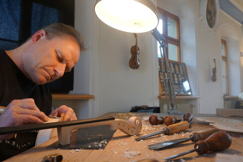 Man blickt auf einen Mann, der mit einem Schnitzmesser in der Hand am Corpus einer Geige arbeitet. Vor ihm auf dem hölzernen Tisch liegt der rohe Geigenhals und weitere Schnitzmesser. Über dem Mann leuchtet eine helle Lampe. Der Mann wirkt konzentriert und fokussiert. 