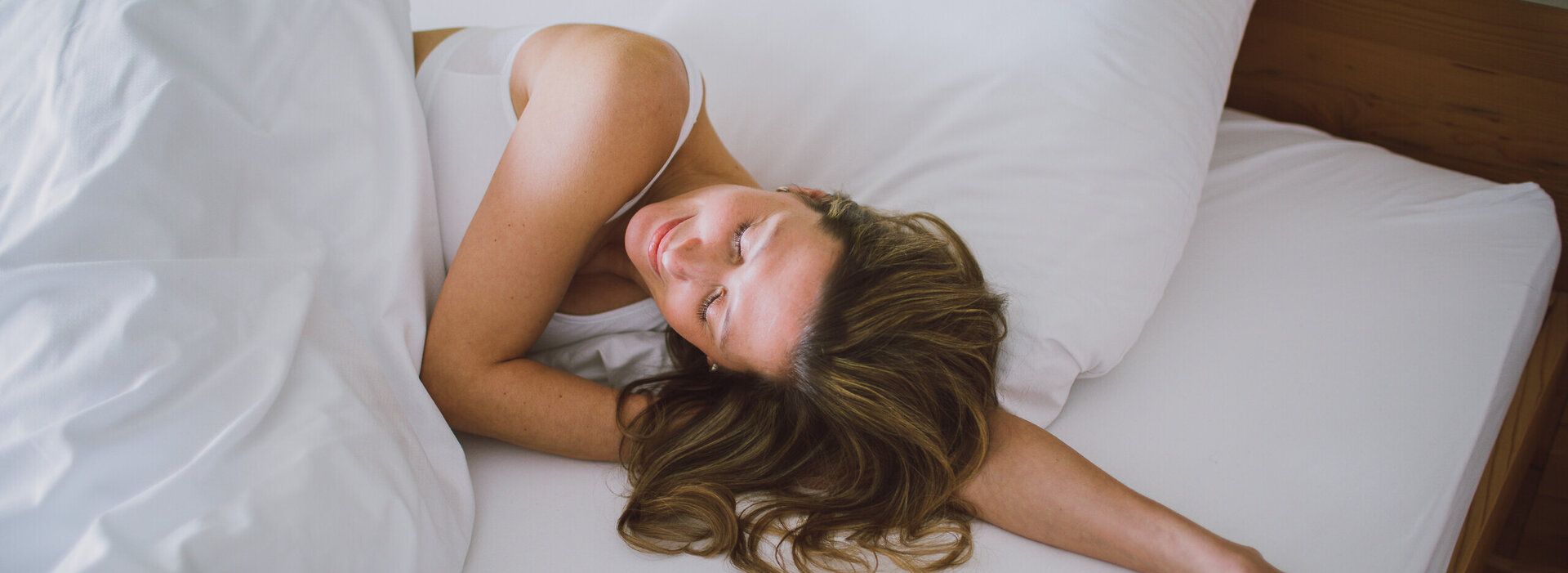 Das Bild zeigt eine Frau mit einem weißen kurzen Oberteil. Sie liegt in einem Bett, das weiß bezogen ist. Ihr Kopf liegt auf ihrem linken Arm. Sie hat die Augen geschlossen und wirkt entspannt. 