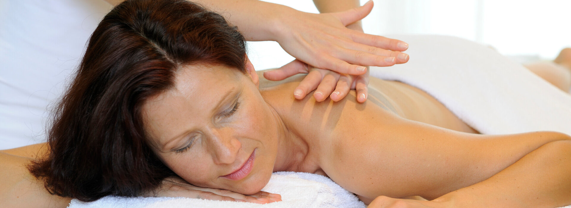 Das Bild zeigt eine Frau, die bäuchlings auf einer Massageliege liegt. Sie ist unbekleidet. Ein weißes Handtuch bedeckt ihr Gesäß und ihren unteren Rücken. Sie hat die Augen geschlossen. Über ihr steht eine Person, die ihr die obere Rückenpartie massiert. Die Person trägt ein weißes Oberteil. Der Raum ist hell erleuchtet.      