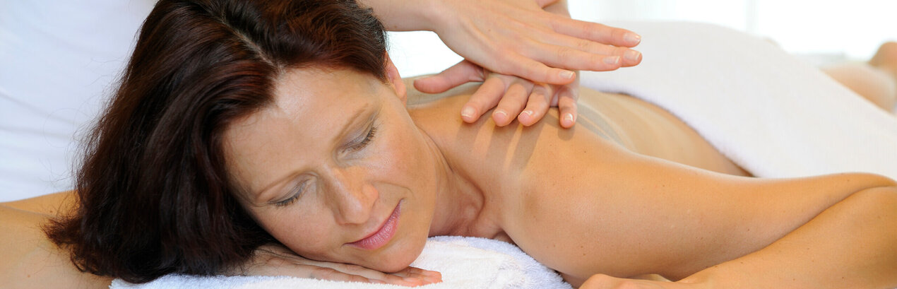 Das Bild zeigt eine Frau, die bäuchlings auf einer Massageliege liegt. Sie ist unbekleidet. Ein weißes Handtuch bedeckt ihr Gesäß und ihren unteren Rücken. Sie hat die Augen geschlossen. Über ihr steht eine Person, die ihr die obere Rückenpartie massiert. Die Person trägt ein weißes Oberteil. Der Raum ist hell erleuchtet.      