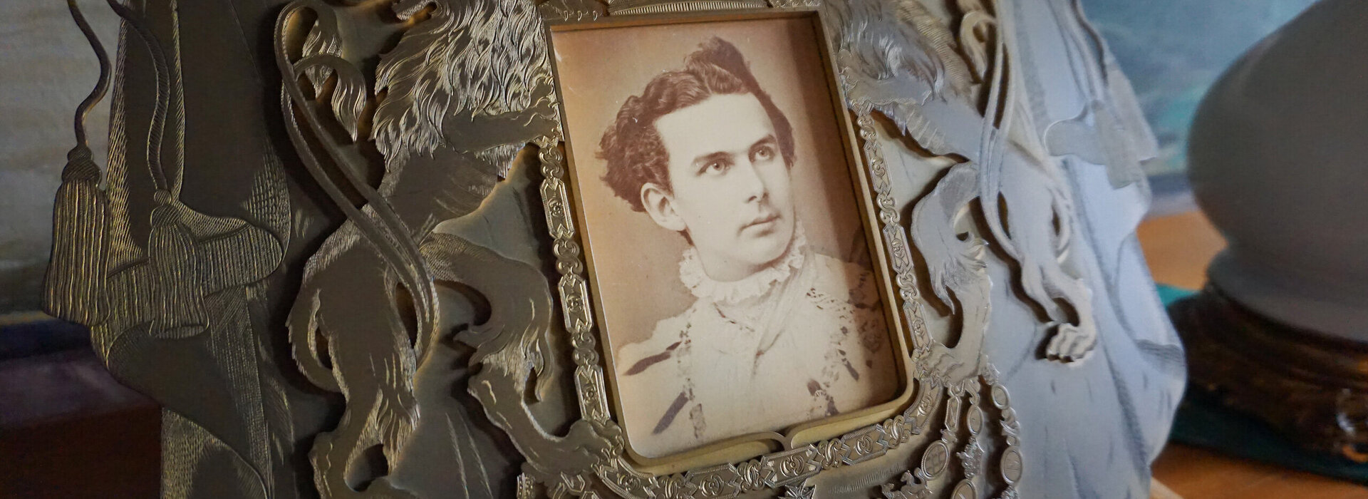 Das Bild zeigt ein Schwarz/weiß Foto von König Ludwig, welches liebevoll in eine verziehrte und geschnitze Holztafel eingebetet wurde. 