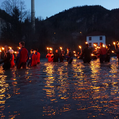 Das Bild zeigt mehrere Personen in Neoprenanzügen im knietiefen Wasser stehen. Alle tragen unterschiedliche Mützen und halten brennende Fackeln in den Händen. Es Dämmert. Es ist schon fast dunkel.  