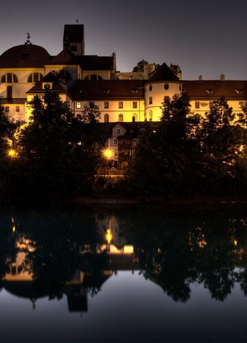 Das Foto zeigt den weitläufigen Gebäudekomplex des ehemaligen Benediktinerklosters St. Mang mit seiner weißen Fassade, roten Dächern und schier unzähligen Fenstern. Es ist Abend. Die Außenbeleuchtung taucht die Szene in ein warmes Licht. Im Vordergrund fließt der Fluß Lech scheinbar bewegungslos. Die Wasseroberfläche ist glatt wie ein Spiegel, in dem sich das Kloster spiegelt.