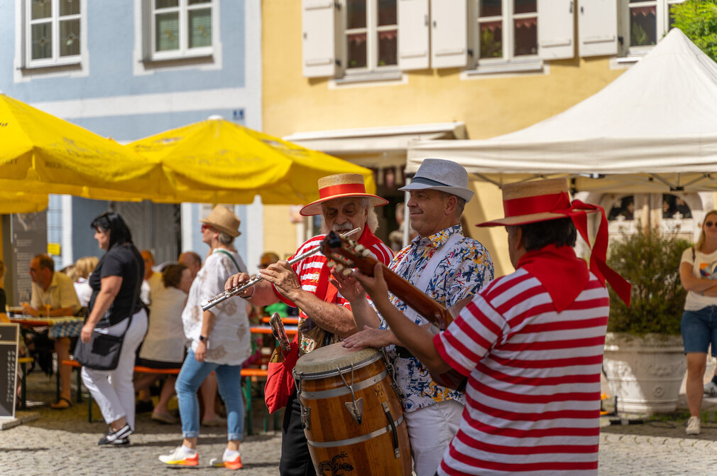 Das Bild zeigt drei Musiker. Ein Musiker spielt Querflöte, einer Gitarre und einer Trommel. Die beiden äußeren Musiker tragen rot-weiße T-Shirts und Strohhüte mit roter Borte. Der Mann in der Mitte trägt ein buntes Hemd, einen hellgrauen Hut und weiße Hosenträger. Im Hintergrund sind Häuserfassaden zu erkennen. Menschen sitzen an Tischen und unterhalten sich. Die Sonne scheint.  