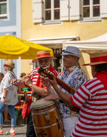 Das Bild zeigt drei Musiker. Ein Musiker spielt Querflöte, einer Gitarre und einer Trommel. Die beiden äußeren Musiker tragen rot-weiße T-Shirts und Strohhüte mit roter Borte. Der Mann in der Mitte trägt ein buntes Hemd, einen hellgrauen Hut und weiße Hosenträger. Im Hintergrund sind Häuserfassaden zu erkennen. Menschen sitzen an Tischen und unterhalten sich. Die Sonne scheint.  