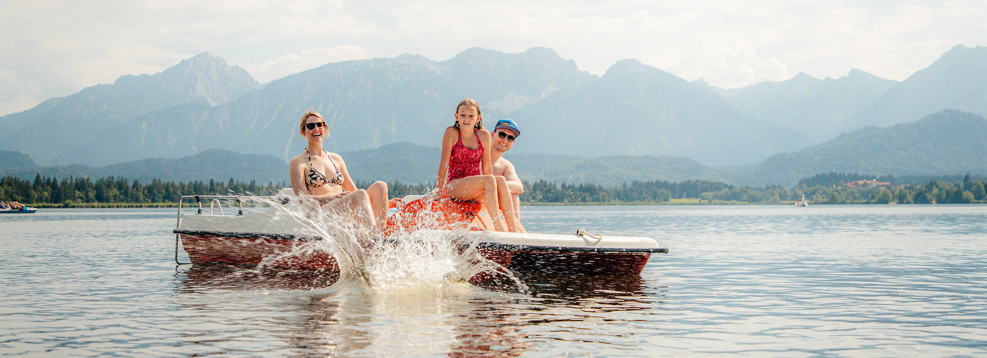 Das Bild zeigt eine idyllische Szene auf einem See, mit drei Personen, die eine Bootsfahrt genießen. Das Boot ist in einem lebhaften Rot lackiert und verfügt über ein weißes Dach, das Schutz für die Passagiere bietet. Das Boot bewegt sich, was durch die aufgewühlten Wasser hinweist, dass es mit einer mäßigen Geschwindigkeit unterwegs ist.  Die drei Personen auf dem Boot sind alle mit Badeanzügen bekleidet, was darauf hinweist, dass sie bereit sind, im See zu schwimmen. Die Person auf der linken Seite sitzt, während die anderen beiden stehen, alle Gesichter zur Kamera gedreht, deren Lächeln Freude und Entspannung ausstrahlen.  Die Kulisse des Bildes ist ein atemberaubender Blick auf Berge, was ein Gefühl von Abenteuer und Entdeckung zur Szene hinzufügt. Der Himmel darüber ist ein klares Blau, mit flauschigen weißen Wolken, die das malerische Setting vervollständigen. Das Gesamtbild erzeugt ein Bild eines perfekten Tages am Wasser, umgeben von der Schönheit der Natur.