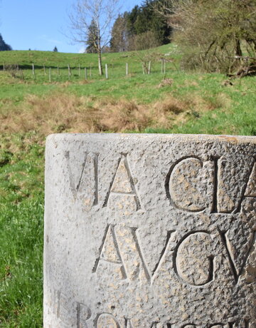 Im Vordergrund des Bildes steht eine Steinsäule, auf der „Via Claudia Augusta“ zu lesen ist. Im Hintergrund ist ein mit grünem Gras bewachsener Hügel zu sehen. Vereinzelt wächst auch Buschwerk und Bäume auf dem Hügel. Mitten durch den Hügel ist ein Zaun gespannt. 