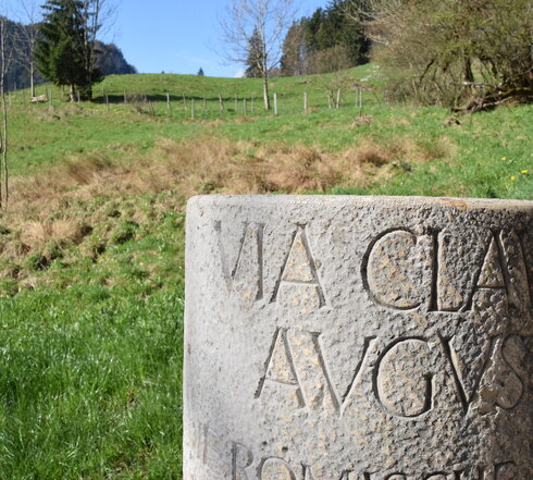 Im Vordergrund des Bildes steht eine Steinsäule, auf der „Via Claudia Augusta“ zu lesen ist. Im Hintergrund ist ein mit grünem Gras bewachsener Hügel zu sehen. Vereinzelt wächst auch Buschwerk und Bäume auf dem Hügel. Mitten durch den Hügel ist ein Zaun gespannt. 