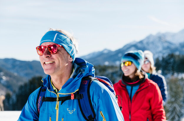 Das Bild zeigt drei Personen in sportlicher Winterkleidung mit Sonnenbrillen und Mützen. Sie laufen hintereinander. Im Hintergrund erheben sich bewaldete, schneebedeckte Berge. Der Himmel ist blau. Die Sonne scheint. Ein Mann läuft voraus. Zwei Frau laufen ihm nach. Alle lächeln. Man kann nur ihre Oberköper und Köpfe sehen. 