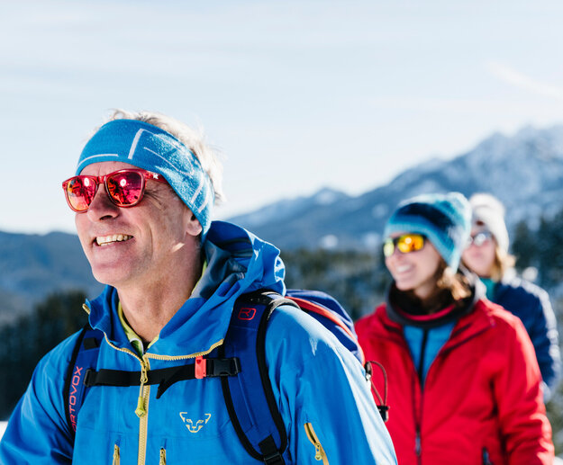Das Bild zeigt drei Personen in sportlicher Winterkleidung mit Sonnenbrillen und Mützen. Sie laufen hintereinander. Im Hintergrund erheben sich bewaldete, schneebedeckte Berge. Der Himmel ist blau. Die Sonne scheint. Ein Mann läuft voraus. Zwei Frau laufen ihm nach. Alle lächeln. Man kann nur ihre Oberköper und Köpfe sehen. 