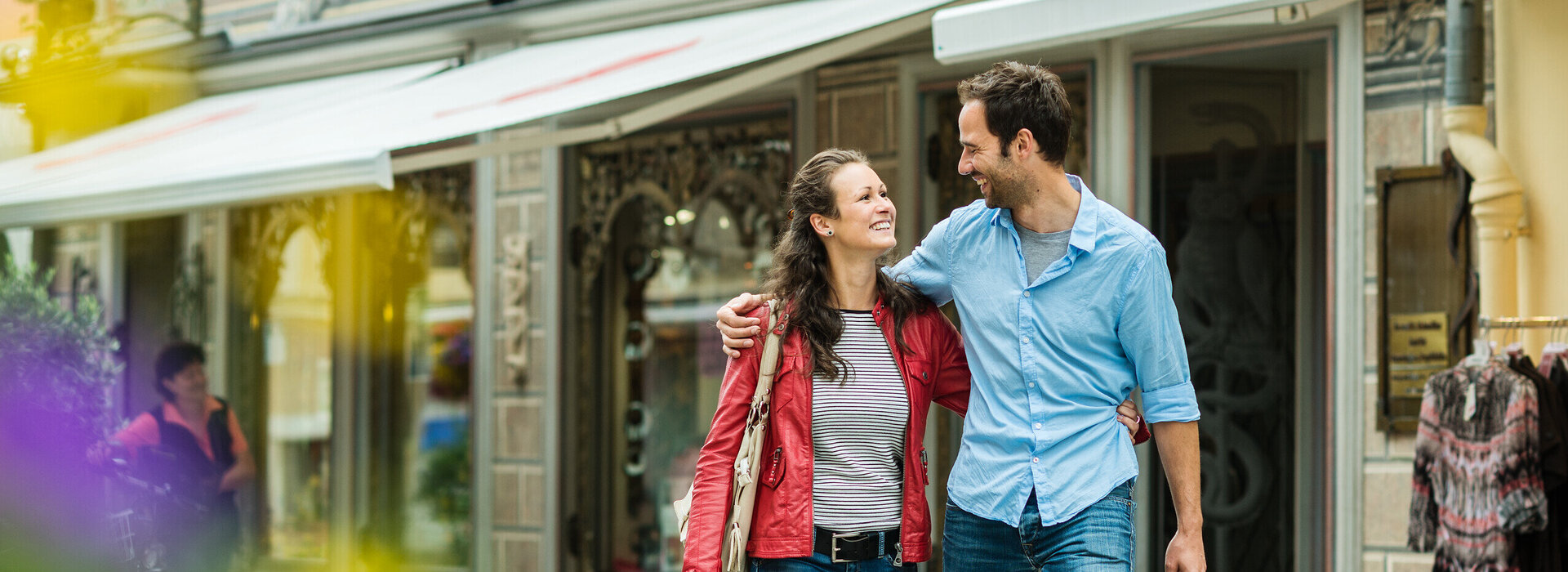 Das Bild zeigt einen jungen Mann und eine Frau. Der Mann hat den Arm um die Frau gelegt. Sie lächeln sich an. Die beiden stehen vor einem reich verzierten Gebäude. Im Hintergrund sind Schaufenster zu sehen. Es ist Sommer. 