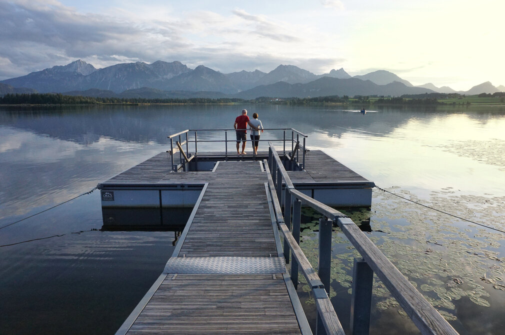 Das Bild fängt eine harmonische Szene auf einer Mole ein. Hierbei handelt es sich um die Kneippinsel in Hopfen am See. Zwei Personen, ein Mann und eine Frau, stehen auf der hölzernen Insel, die von einem Metallgeländer zum Wasser hin begrenzt ist. Die Insel befindet sich auf dem Hopfensee in Ufernähe und ist mit einem Holzsteg mit der Uferpromenade verbunden. In der Ferne erheben sich majestätische Berge gegen den Himmel, was der Szene eine Atmosphäre der Ruhe und Gelassenheit verleiht. Der Mann und die Frau scheinen die Aussicht zu genießen, vielleicht eine Pause von ihrem Tag zu machen oder einfach die Schönheit der Natur zu schätzen. Die allgemeine Atmosphäre des Bildes ist friedvoll und beschaulich, was ein Gefühl von Ruhe und Entspannendes hervorruft.