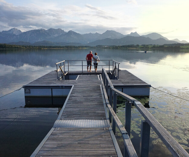 Das Bild fängt eine harmonische Szene auf einer Mole ein. Hierbei handelt es sich um die Kneippinsel in Hopfen am See. Zwei Personen, ein Mann und eine Frau, stehen auf der hölzernen Insel, die von einem Metallgeländer zum Wasser hin begrenzt ist. Die Insel befindet sich auf dem Hopfensee in Ufernähe und ist mit einem Holzsteg mit der Uferpromenade verbunden. In der Ferne erheben sich majestätische Berge gegen den Himmel, was der Szene eine Atmosphäre der Ruhe und Gelassenheit verleiht. Der Mann und die Frau scheinen die Aussicht zu genießen, vielleicht eine Pause von ihrem Tag zu machen oder einfach die Schönheit der Natur zu schätzen. Die allgemeine Atmosphäre des Bildes ist friedvoll und beschaulich, was ein Gefühl von Ruhe und Entspannendes hervorruft.