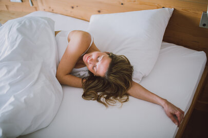 Das Bild zeigt eine Frau mit einem weißen kurzen Oberteil. Sie liegt in einem Bett, das weiß bezogen ist. Ihr Kopf liegt auf ihrem linken Arm. Sie hat die Augen geschlossen und wirkt entspannt. 