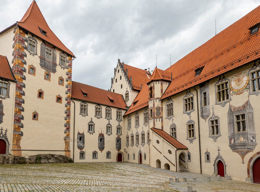 Das Bild zeigt ein große mit Illusionsmalerei verziertes Gebäude. Es hat viele Fenster und rote Dächer. Der Gebäudekomplexes besteht aus mehreren unterschiedlichen Gebäudeteilen, die aneinandergebaut sind. Der Himmel darüber ist wolkenverhangen.