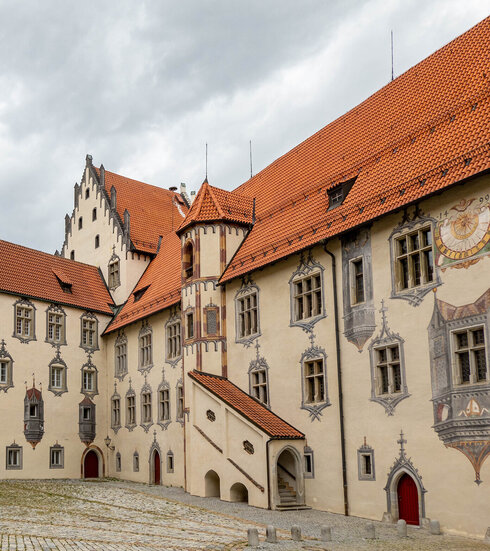 Das Bild zeigt ein große mit Illusionsmalerei verziertes Gebäude. Es hat viele Fenster und rote Dächer. Der Gebäudekomplexes besteht aus mehreren unterschiedlichen Gebäudeteilen, die aneinandergebaut sind. Der Himmel darüber ist wolkenverhangen.