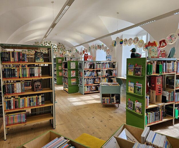 Auf dem Bild ist der Innenraum einer Bibliothek zu sehen. Die Regale sind bunt und mit Büchern gefüllt. 