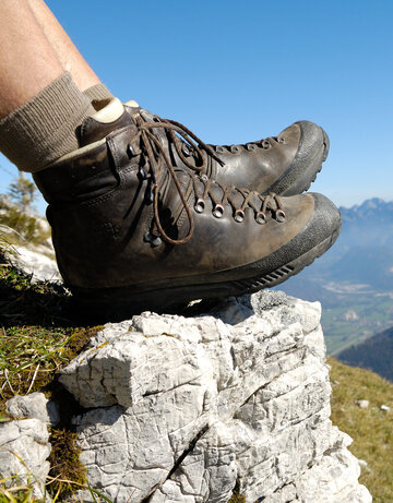 Das Bild zeigt eine Person, die auf einem Berg sitzt und nach unten blickt. Allerdings sind nur die ledernen Bergschuhe und die Wadern der Person zu sehen. Weiter unten erkennt man Bergwiesen, Wälder und ein Tal. Dahinter erheben sich weitere Berge. Der Himmel ist blau. Die Sonne scheint.  